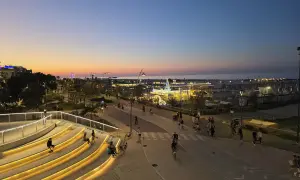 Wieczorny spacer po promenadzie w Riccione. Ludzie odpoczywają na podświetlonych schodach, inni jeżdżą na rowerach. W oddali widać morze i wesołe miasteczko.