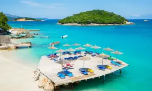 Słoneczny dzień na Ksamil Beach. Drewniany taras z leżakami i parasolami, turkusowa woda i zielona wyspa w tle.