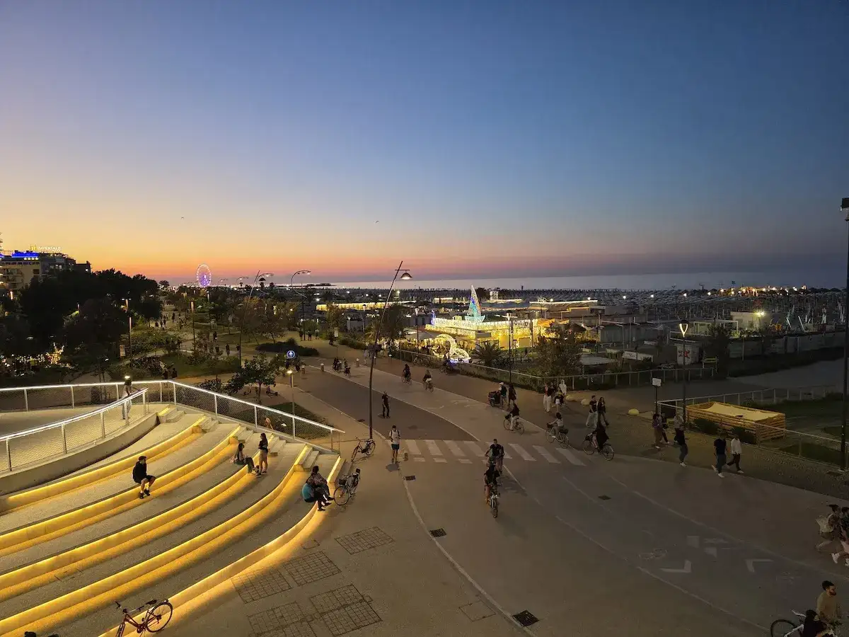 Wieczorny spacer po promenadzie w Riccione. Ludzie odpoczywają na podświetlonych schodach, inni jeżdżą na rowerach. W oddali widać morze i wesołe miasteczko.