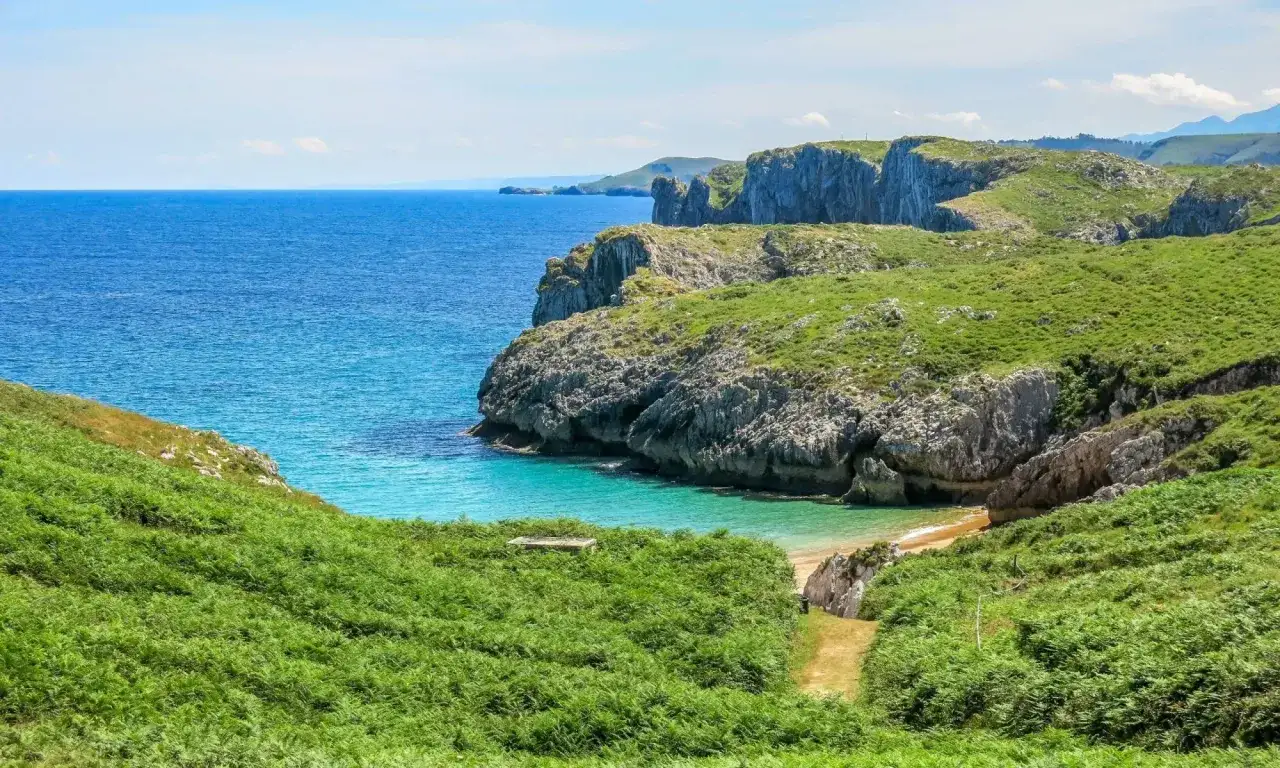 Zielone wzgórza i klify nad turkusowym morzem. Ścieżka prowadzi w dół do ukrytej plaży, idealnej na odpoczynek po długim camino.
