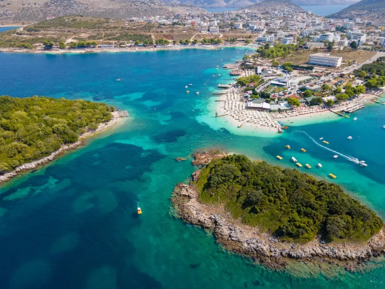 Malowniczy widok na Ksamil Beach z turkusową wodą, zielonymi wyspami i zatłoczoną plażą pełną leżaków.