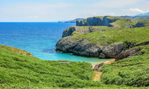 Zielone wzgórza i klify nad turkusowym morzem. Ścieżka prowadzi w dół do ukrytej plaży, idealnej na odpoczynek po długim camino.